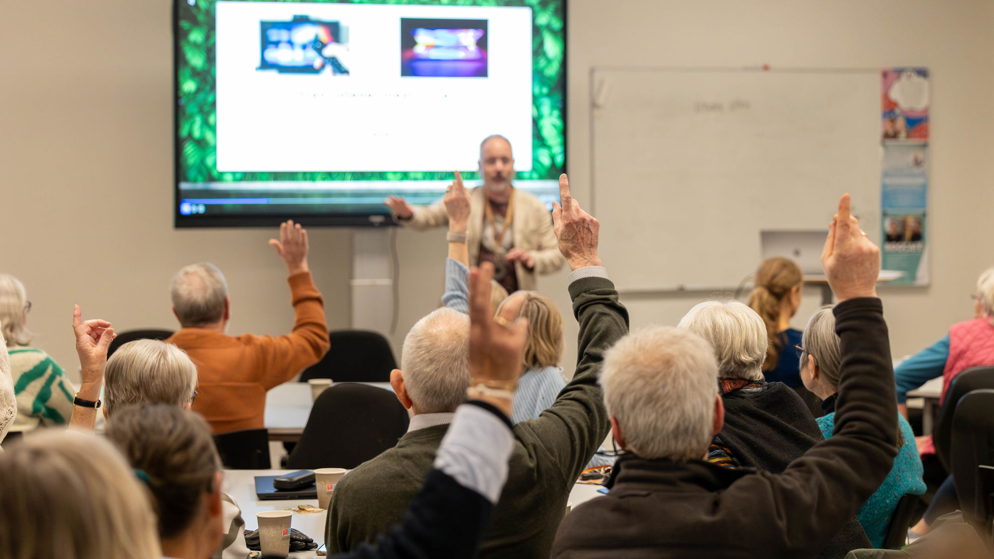 Møde. En mand står med fronten mod kamera og deltagere sidder med ryggen til og rækker hånden op